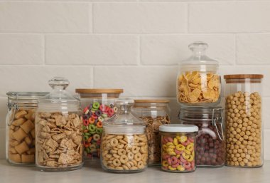 Tuğla duvarın yanındaki beyaz tezgahta farklı kahvaltı gevrekleri olan cam kaplar.