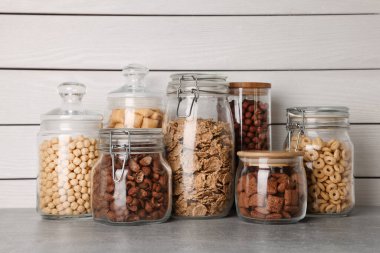 Glass containers with different breakfast cereals on grey countertop near white wooden wall