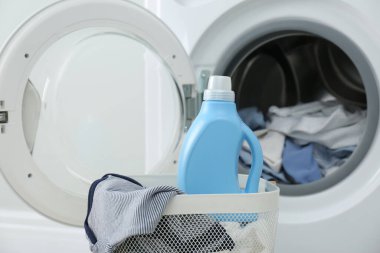 Çamaşır makinesinin yanındaki sepette bir şişe deterjan ve çocuk giysileri var.