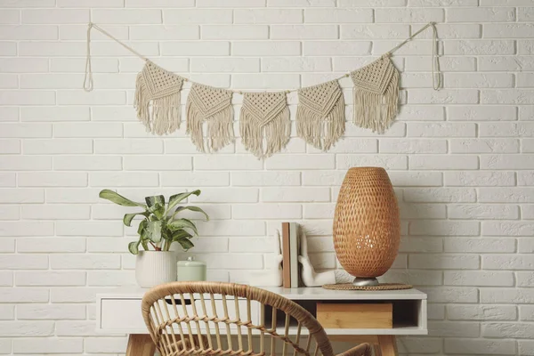 Potted houseplant, decor elements and books on white table near brick wall with stylish macrame