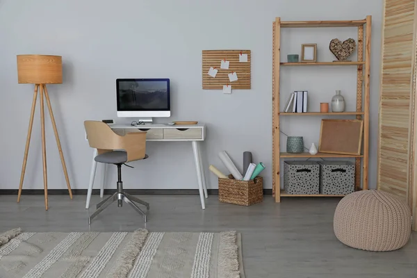 Stylish room interior with comfortable workplace and shelving unit near white wall