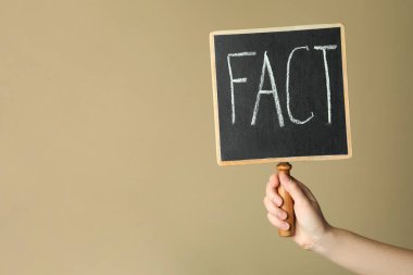 Woman holding small chalkboard with word FACT near beige wall, closeup. Space for text