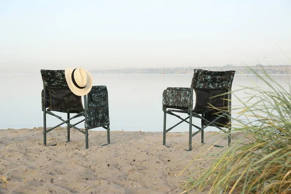 Camouflage fishing chairs with hat on sandy beach near river