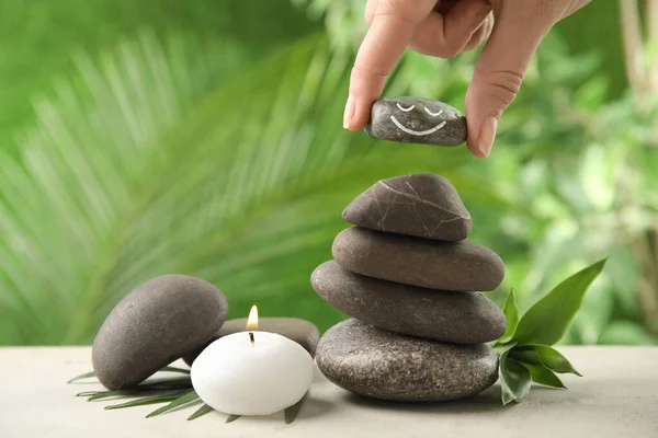 Woman putting stone with drawn happy face on stack against blurred green background, closeup. Be in harmony and enjoying your life