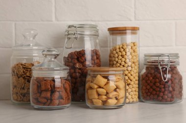 Tuğla duvarın yanındaki beyaz tezgahta farklı kahvaltı gevrekleri olan cam kaplar.