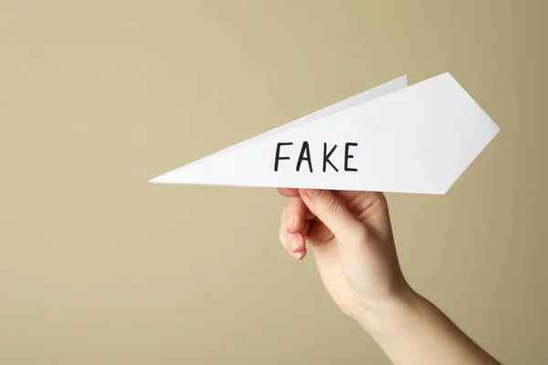 Woman holding paper plane with word FAKE near beige wall, closeup