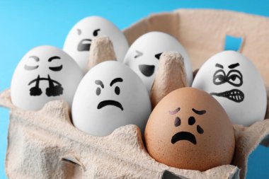 Brown egg with upset face among aggressively disposed white ones in carton box on turquoise background, closeup. Bullying concept