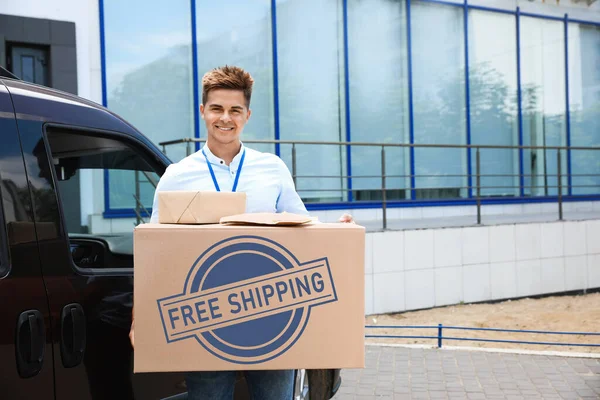 Young courier holding parcels near delivery car outdoors. Free shipping