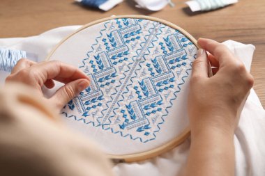 Woman embroidering white shirt with blue thread at wooden table, closeup. Ukrainian national clothes