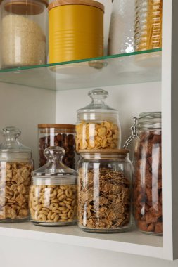 Farklı kahvaltı gevrekleri ve raflardaki diğer ürünlerle dolu cam kaplar.