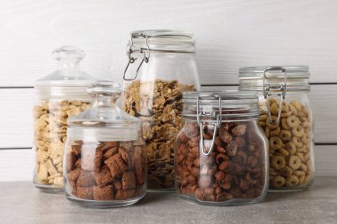 Glass containers with different breakfast cereals on grey countertop near white wooden wall