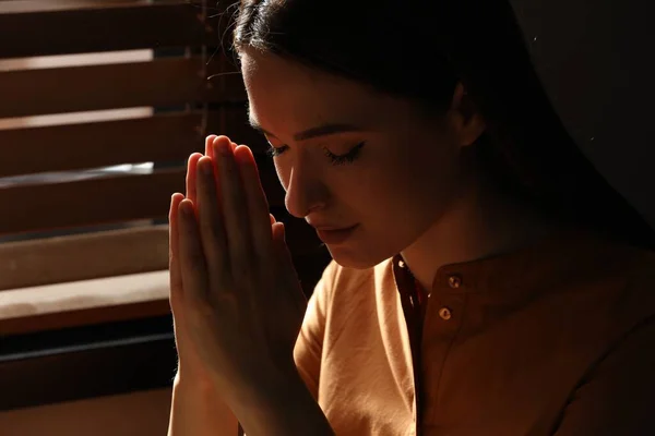 Religious young woman with clasped hands praying indoors, closeup