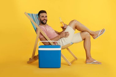 Happy man with bottle of beer resting in deck chair near cool box on yellow background