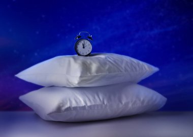 Soft pillows and alarm clock on light grey table against night sky with stars. Insomnia