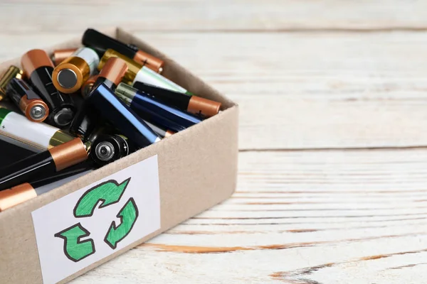 Used batteries in cardboard box with recycling symbol on white wooden table, closeup. Space for text