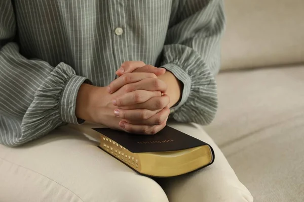 Religious woman praying over Bible on sofa, closeup