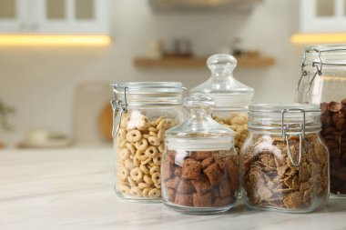 Glass containers with different breakfast cereals on white marble table in kitchen. Space for text