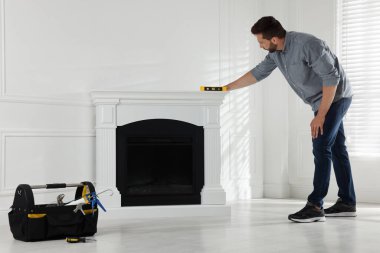Man using construction level for installing electric fireplace near white wall in room