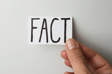 Woman holding sheet of paper with word Fact on light grey background, top view
