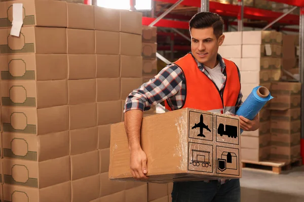 Worker holding roll of stretch film and wrapped box with shipping icons in warehouse