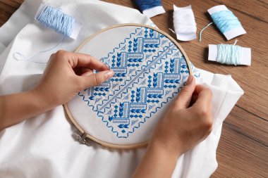 Woman embroidering white shirt with blue thread at wooden table, above view. Ukrainian national clothes