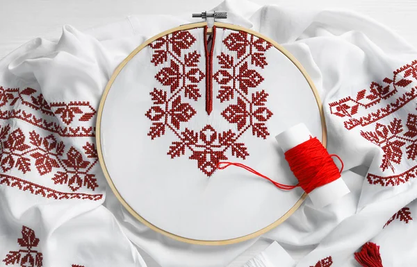 Shirt with red embroidery design in hoop, needle and thread on table, top view. National Ukrainian clothes
