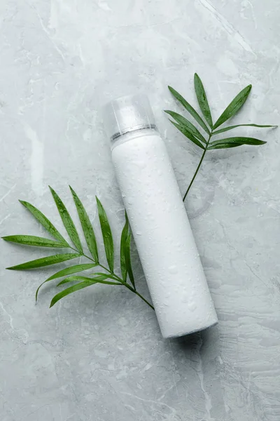 Dry shampoo spray and green leaves on light grey table, flat lay