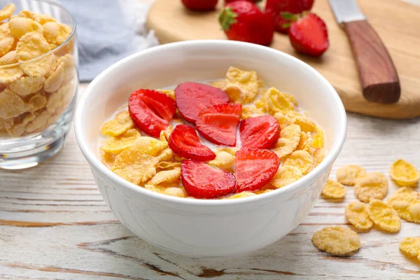 Bowl of tasty crispy corn flakes with milk and strawberries on white wooden table