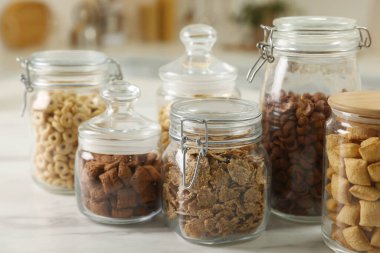 Glass containers with different breakfast cereals on white marble table indoors