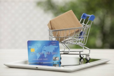 Online payment concept. Small shopping cart with bank card, box and tablet on white table, closeup