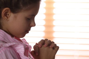 Cute little girl with hands clasped together praying near window, closeup. Space for text