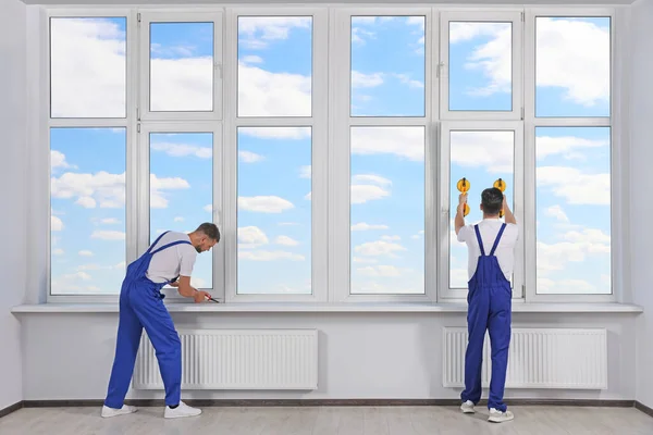 Workers with tube of sealant and suction lifters installing plastic windows indoors