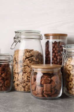 Glass containers with different breakfast cereals on grey countertop near white wooden wall