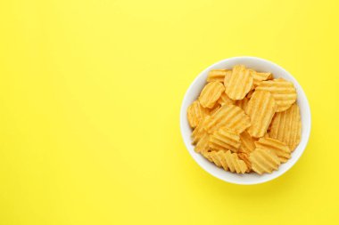 Bowl of tasty ridged potato chips on yellow background, top view. Space for text