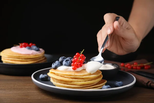 Woman putting natural yogurt onto tasty pancakes with blueberries and red currants at wooden table, closeup. Space for text
