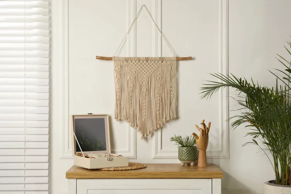 Open jewelry box and decor elements on chest of drawers near white wall with stylish macrame