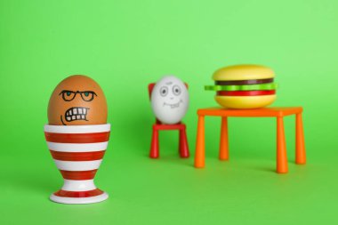 Angry egg and happy one with burger at table on green background