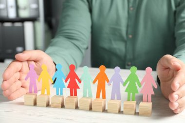 Man protecting paper human figures with wooden cubes at table indoors, closeup. Diversity and Inclusion concept