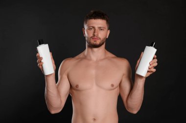 Shirtless young man holding bottles of shampoo on black background