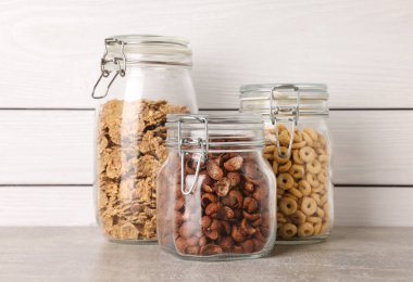 Glass containers with different breakfast cereals on grey countertop near white wooden wall