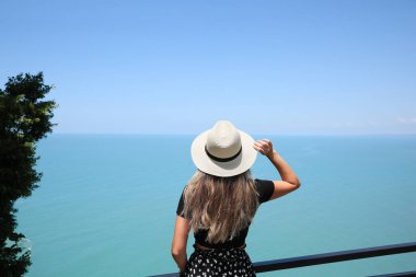 Woman with hat near sea on sunny day, back view