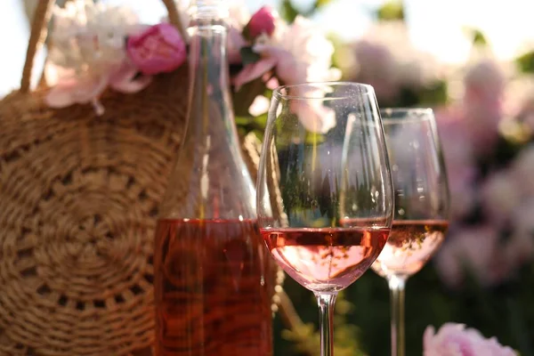 Bottle and glasses of rose wine near straw bag with beautiful peonies in garden, closeup