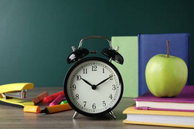 Alarm saati ve yeşil karatahtanın yanındaki ahşap masada farklı kırtasiye malzemeleri. Okul zamanı.