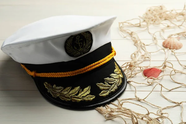 Peaked cap, net and shells on white wooden background, space for text