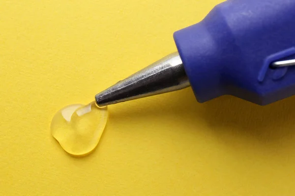 Melted glue dripping out of hot gun nozzle on yellow background, closeup