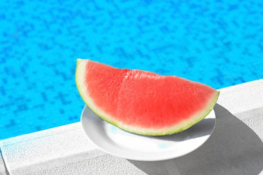 Slice of fresh juicy watermelon on white plate near swimming pool outdoors