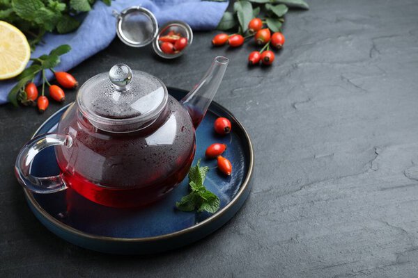 Teapot with aromatic rose hip tea, berries and mint on black table, space for text