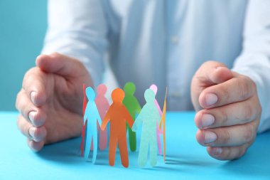 Man protecting paper human figures on light blue table, closeup. Diversity and inclusion concept