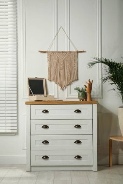 Cozy room interior with chest of drawers and stylish macrame on wall