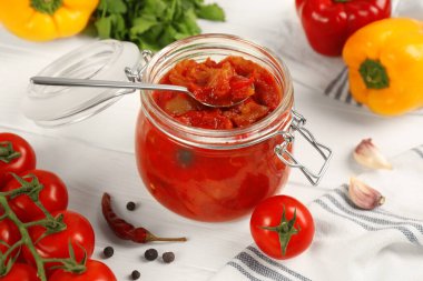 Glass jar of delicious canned lecho and fresh ingredients on white wooden table, closeup